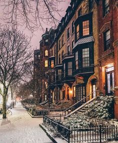 a snow covered sidewalk next to tall brick buildings and trees with lights on at night