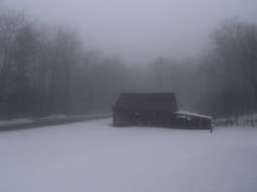 a barn in the middle of a snow covered field with trees and bushes behind it