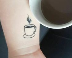 a cup of coffee is on the wrist of a person with a small tattoo design