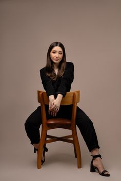 a woman sitting on top of a wooden chair in front of a gray background with her legs crossed