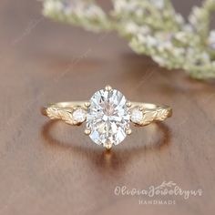 a gold ring with an oval cut diamond in the center on a wooden table next to flowers