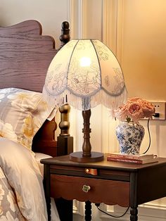 a lamp on a night stand next to a bed with pillows and flowers in vases