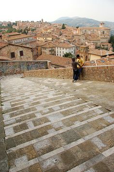 two people are sitting on a stone wall overlooking the city and surrounding it, while another person is looking at something in the distance