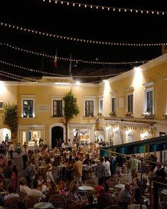 people sitting at tables in front of a building with lights strung over it and on the ground