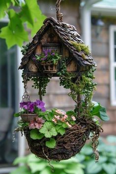 a bird house hanging from a tree branch filled with flowers and greenery in front of a house