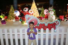 a little boy standing in front of a fence with christmas decorations on it and snowmen behind him