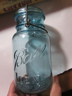 a hand holding a blue glass jar with writing on the top and bottom, in front of a book