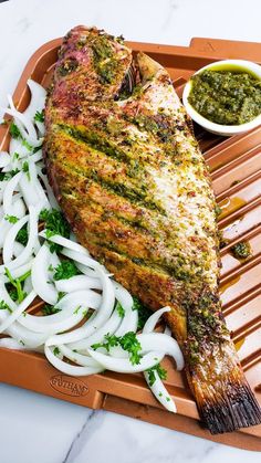 grilled fish with onions and green sauce on a tray next to a side dish