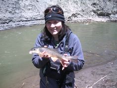 a woman holding a fish in her hands while standing next to a river with rocks and water