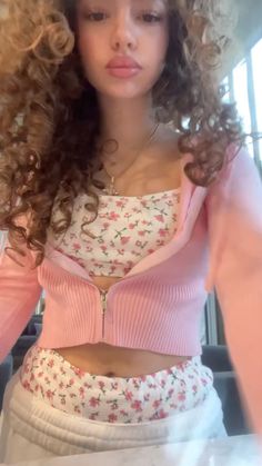 a woman with curly hair wearing a pink sweater and white skirt is looking at the camera