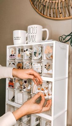 two hands reaching for magnets on top of a white cabinet with drawers and mugs