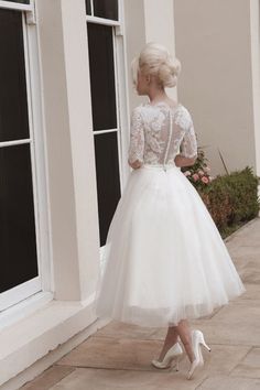 a woman in a white dress is standing near a window and looking at the ground