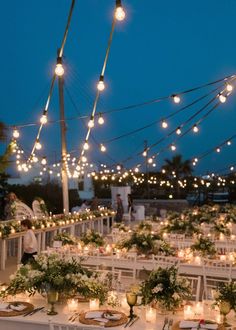 an outdoor dining area with white tables and lights strung from the ceiling over them, surrounded by greenery