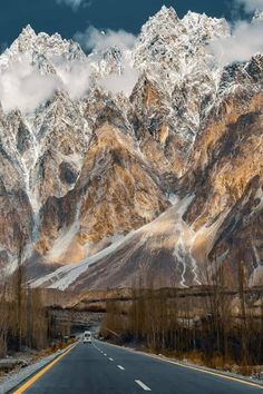 the mountains are covered with snow and clouds in the distance is an empty road surrounded by bare trees