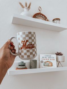 a person holding a coffee mug with the word mama on it in front of a shelf