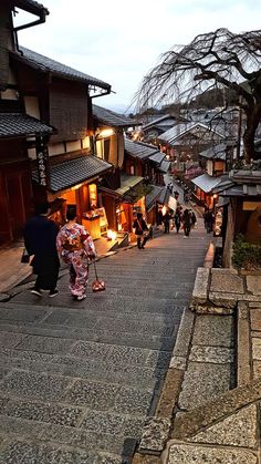 a woman walking down a street next to tall buildings with shops on each side and trees in the background