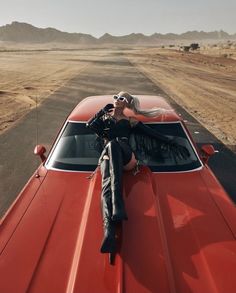 a woman sitting on the hood of a red car in the middle of an empty road