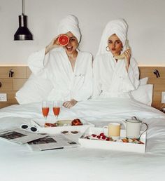 two women in bathrobes sitting on a bed with food and drinks next to them