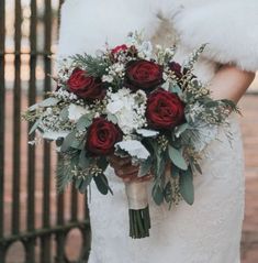 a bride holding a bouquet of red roses and white carnations in front of an iron gate