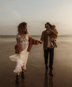 a man and woman walking on the beach with a baby in their arms while holding hands