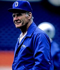 an older man wearing a baseball cap and blue jacket standing on a field with other players in the background