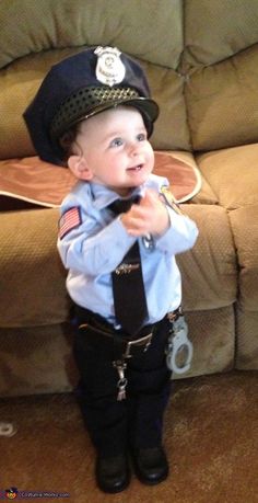 a little boy dressed up as a police officer standing in front of a couch wearing a tie and hat