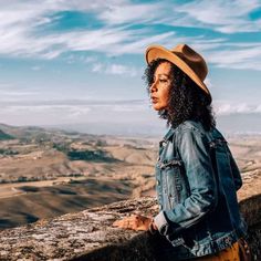 a woman with curly hair wearing a denim jacket and hat standing on top of a hill