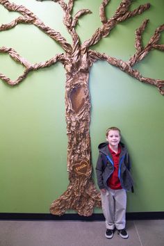 a young boy standing in front of a tree made out of paper mache and cardboard