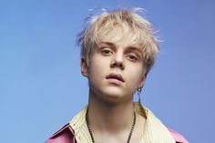 a young man with blonde hair wearing a necklace and looking off to the side, against a blue background