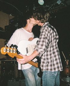 two young men are playing guitars together in a music studio, one is kissing the other's cheek