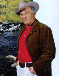 an old man wearing a cowboy hat and standing in front of a river with rocks