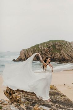 a woman in a white dress is standing on the rocks by the ocean with her arms outstretched