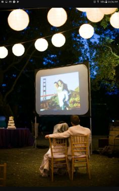 two people sitting at a table watching a movie on the screen with paper lanterns hanging above them