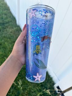 a hand holding a blue mermaid tumbler with stars and fish on the bottom, in front of a white fence