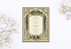 an ornate wedding card on top of a table next to some white flowers and greenery