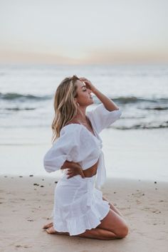 a woman sitting on top of a sandy beach next to the ocean wearing a white dress