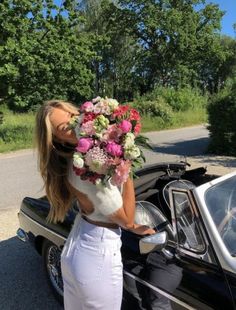 a woman in white pants holding a bouquet of flowers next to a black car