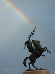 a statue with a rainbow in the sky behind it and a person on a horse holding a spear