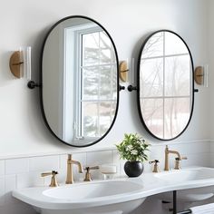 two oval mirrors are hanging on the wall above double sinks in a white bathroom with gold faucets