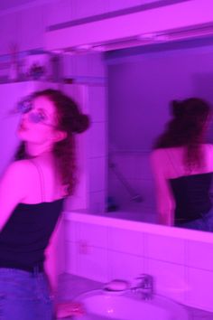 a woman standing in front of a bathroom mirror next to a white sink under a purple light