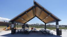 an outdoor picnic area with tables and benches under a wooden roof on a sunny day