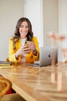 a woman sitting at a table looking at her cell phone and smiling while she looks at her laptop