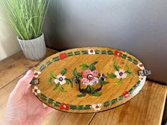 a hand holding a decorative wooden tray with flowers on it and a potted plant in the background