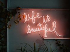 a neon sign that says life is beautiful on the side of a wall next to plants