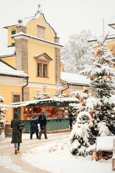 two people are walking in the snow near a christmas tree and small yellow building with a clock on it