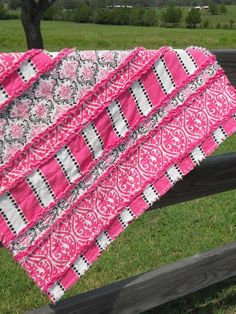 a pink and black quilt on a wooden fence in front of a field with trees