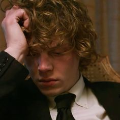 a young man with curly hair wearing a suit and tie, holding his hand to his head