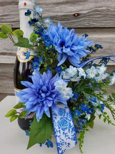 blue flowers and greenery sit in front of a bottle of wine on a table