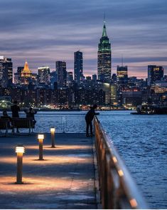 people are sitting on benches near the water at night in front of city lights and skyscrapers