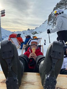 a person sitting on top of a wooden bench eating food next to skis and snow covered mountains